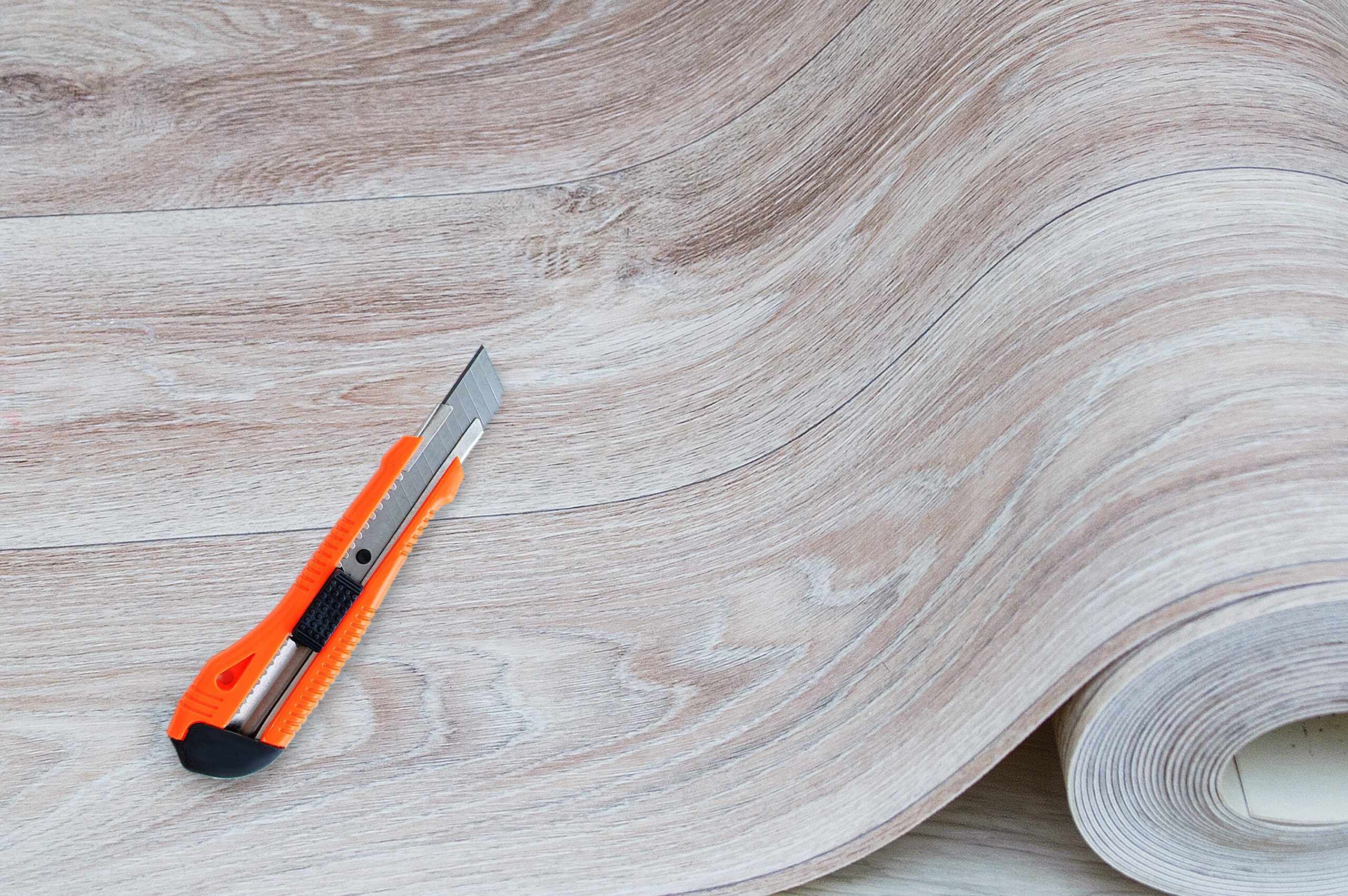 Unwound roll of beige linoleum with wood texture and construction knife for cutting.